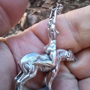 May include: A silver figurine depicting a nude figure riding a canine-like animal. The figurine is detailed, with the rider's arms raised and the animal's posture suggesting movement. The base is a rough, textured shape.