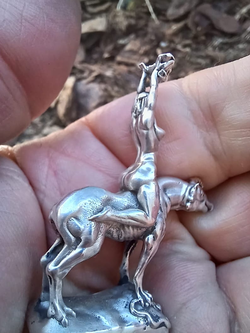 May include: A silver figurine depicting a nude figure riding a canine-like animal. The figurine is detailed, with the rider's arms raised and the animal's posture suggesting movement. The base is a rough, textured shape.
