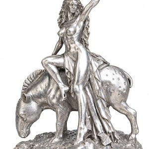 Puede incluir: Una escultura metálica de color plateado que representa una figura de cabello largo y fluido sentada sobre un animal moteado. La figura mantiene un brazo levantado, sosteniendo un ave con las alas extendidas. La base tiene textura de suelo rocoso.