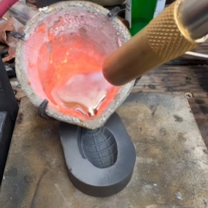 Puede incluir: Primer plano de un proceso de fundición de metal, con metal fundido vertido en un molde con forma de granada. El metal está al rojo vivo y se vierte desde un crisol.