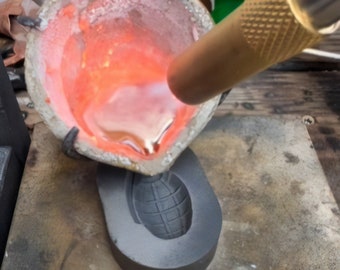 May include: A close-up of a metal casting process, with molten metal being poured into a mold shaped like a grenade. The metal is glowing red hot and is being poured from a crucible.