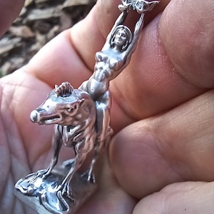 May include: A silver figurine depicting a nude woman riding a wolf-like creature, holding a butterfly aloft. The sculpture is detailed and stands on a rectangular base. The figurine is held in a hand.