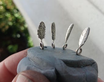 May include: Four small silver-toned metal charms shaped like feathers, each featuring a circular loop at the base. The charms are arranged in a row and are embedded upright into a piece of grey, textured, malleable modeling clay.