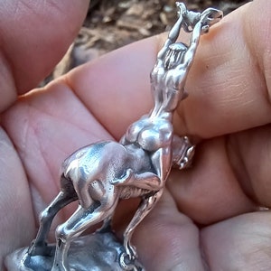 May include: A small, silver sculpture depicting a figure atop an animal, possibly a deer or horse. The figure is reaching upwards, holding an object. The sculpture is detailed and appears to be made of metal, with a polished finish. The base is a rough, rectangular shape.