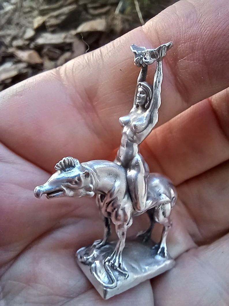 May include: A small, silver figurine depicting a nude figure riding a stylized animal. The figure is holding an object above their head. The sculpture is detailed and appears to be made of metal. The figurine is held in a hand.