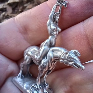 May include: A silver figurine depicting a nude woman riding a mythical beast, holding an object aloft. The sculpture is detailed, with the woman's form and the beast's features clearly defined. The base is rectangular.