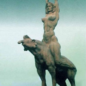 May include: A weathered stone sculpture of a nude woman riding a beast, holding an object aloft. The sculpture is set against a muted teal background. The woman's arms are raised, and the beast has a prominent snout.