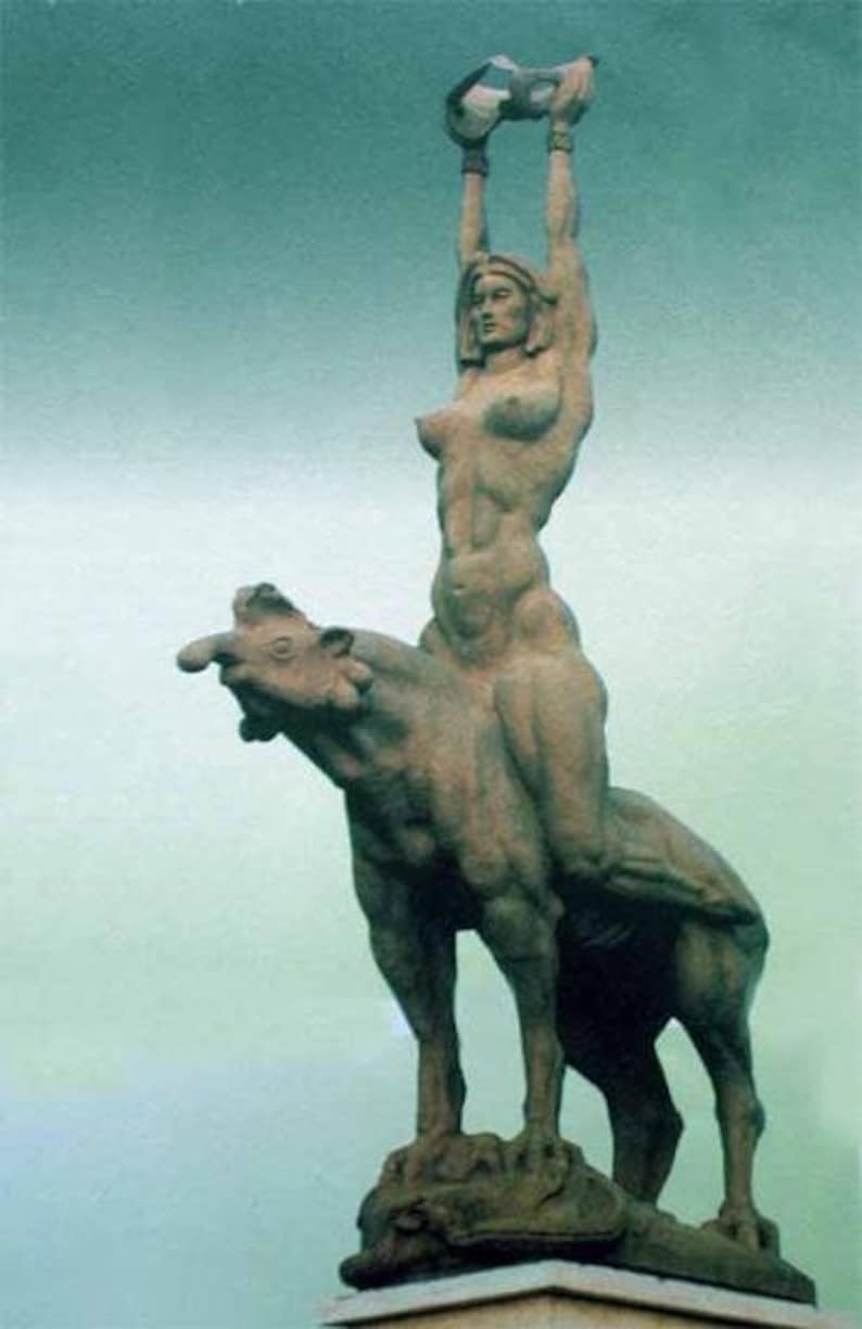 May include: A weathered stone sculpture of a nude woman riding a beast, holding an object aloft. The sculpture is set against a muted teal background. The woman's arms are raised, and the beast has a prominent snout.