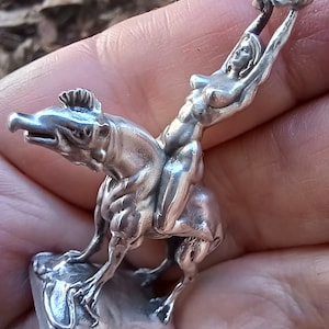 May include: A silver figurine depicting a woman riding a creature with a reptilian head. The woman is reaching upwards, holding an object. The figurine is detailed and appears to be made of metal.