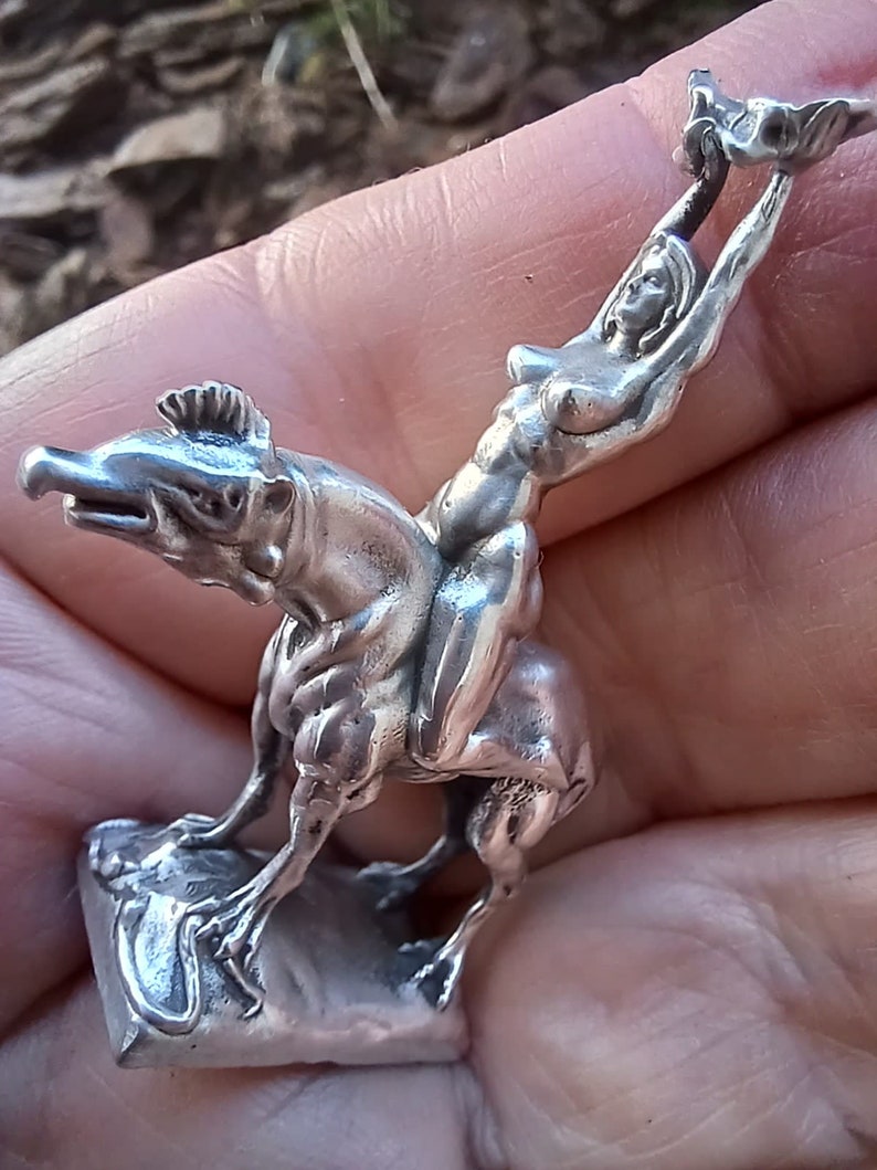 May include: A silver figurine depicting a woman riding a creature with a reptilian head. The woman is reaching upwards, holding an object. The figurine is detailed and appears to be made of metal.