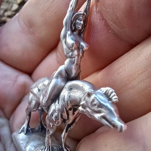 May include: A silver-colored sculpture of a woman riding a dog-like creature, with the woman holding an object above her head. The sculpture is detailed and appears to be made of metal. The base is rectangular.