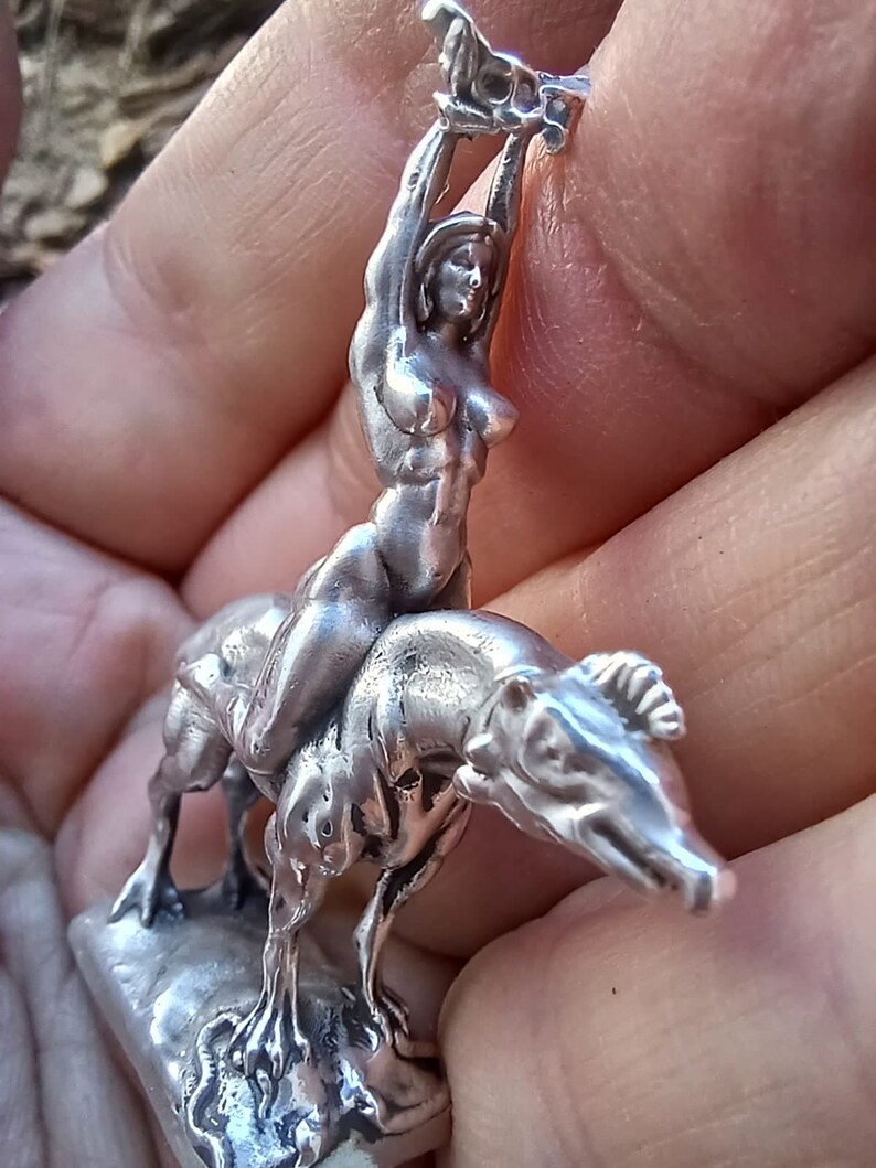 May include: A silver-colored sculpture of a woman riding a dog-like creature, with the woman holding an object above her head. The sculpture is detailed and appears to be made of metal. The base is rectangular.