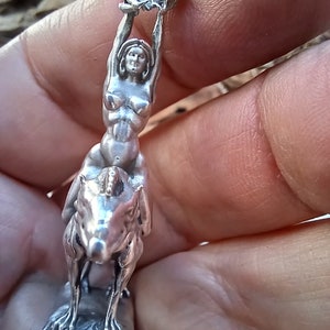May include: A silver figurine depicting a woman riding a horse, holding a decorative object above her head. The figurine is detailed and appears to be made of metal. The woman is nude. The horse is rearing up on its hind legs.