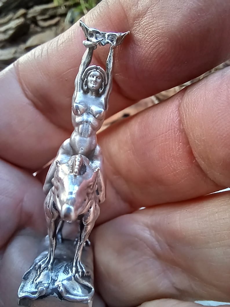 May include: A silver figurine depicting a woman riding a horse, holding a decorative object above her head. The figurine is detailed and appears to be made of metal. The woman is nude. The horse is rearing up on its hind legs.