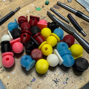 May include: A collection of colorful, 3D-printed components in various shapes and sizes, including spheres, cylinders, and rectangular pieces. The components are in shades of red, black, pink, yellow, blue, and white. Several tools are also visible in the background.