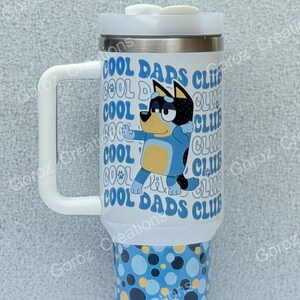 May include: A white stainless steel travel mug with a blue and yellow cartoon dog design. The mug has a white handle and lid. The text "Cool Dads Club" is printed on the mug.