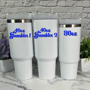 May include: Three white stainless steel tumblers with blue text. The largest tumbler is 40 oz and says "40oz Tumbler 1". The middle tumbler is 40 oz and says "40oz Tumbler 2". The smallest tumbler is 30 oz and says "30oz".