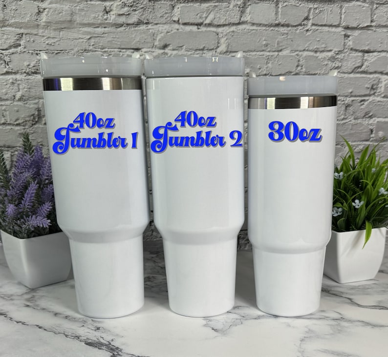May include: Three white stainless steel tumblers with blue text. The largest tumbler is 40 oz and says "40oz Tumbler 1". The middle tumbler is 40 oz and says "40oz Tumbler 2". The smallest tumbler is 30 oz and says "30oz".