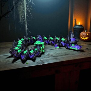 May include: A Halloween-themed, articulated dragon figurine in black, purple, and green. The dragon has a witch hat and cauldron design. The figurine is on a wooden surface with a carved pumpkin and candle in the background.