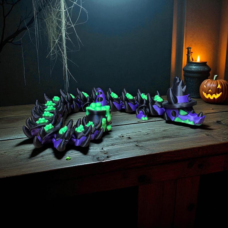 May include: A Halloween-themed, articulated dragon figurine in black, purple, and green. The dragon has a witch hat and cauldron design. The figurine is on a wooden surface with a carved pumpkin and candle in the background.