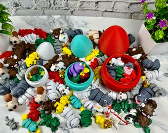 Op de afbeelding: Een verzameling kleurrijke, kleine, plastic speelgoedjes in de vorm van dieren. De speelgoedjes zijn verspreid over een wit oppervlak en sommige bevinden zich in eivormige containers. De speelgoedjes zijn in verschillende kleuren, waaronder rood, blauw, groen, geel en paars.