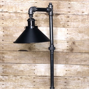 May include: A black industrial style table lamp with a black metal shade and a wooden base. The lamp is made from pipe fittings and has a simple, modern design.