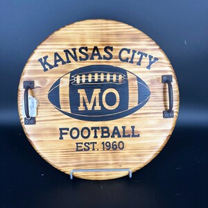May include: A round wooden serving tray with black painted handles. The tray has a black and white football design with the text "KANSAS CITY MO FOOTBALL EST. 1960".
