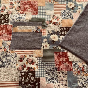 Puede incluir: Una colcha de retazos con una variedad de patrones florales en tonos de azul, rojo y blanco. La colcha est&aacute; hecha de diferentes telas y texturas, incluyendo algod&oacute;n y franela. La colcha est&aacute; bordeada con una tela gris.