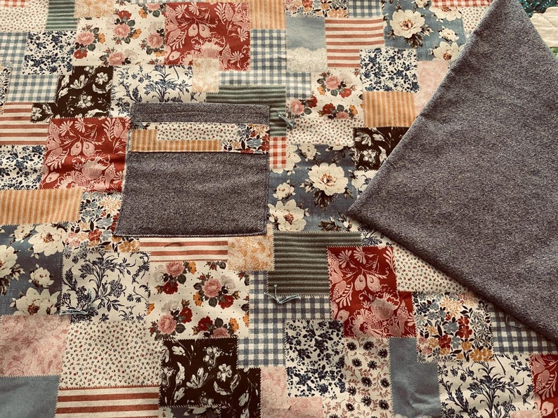 Puede incluir: Una colcha de retazos con una variedad de patrones florales en tonos de azul, rojo y blanco. La colcha est&aacute; hecha de diferentes telas y texturas, incluyendo algod&oacute;n y franela. La colcha est&aacute; bordeada con una tela gris.