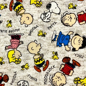 May include: A white fabric with a repeating pattern of cartoon characters. The characters are all from the Peanuts comic strip, including Snoopy, Charlie Brown, Lucy, and Linus. The fabric is printed with the characters' names and phrases like "Snoopy" and "Good ol' Charlie Brown".