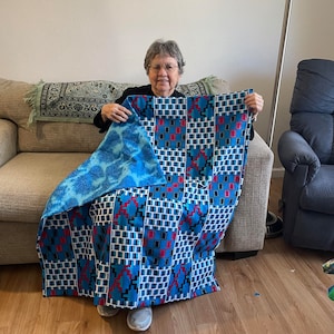 May include: A woman is sitting on a couch holding a blue and red patterned quilt. The quilt has a geometric design with squares and rectangles.