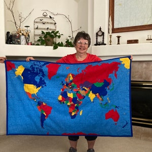 May include: A woman holds up a large world map printed on a blue fabric with a red, yellow, green, and blue color scheme. The map is framed with a dark blue border.