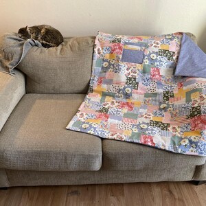 Puede incluir: Un sof&aacute; gris con una colcha de patchwork en tonos azules, rosas, amarillos y verdes. La colcha tiene un patr&oacute;n floral y est&aacute; doblada en el sof&aacute;.