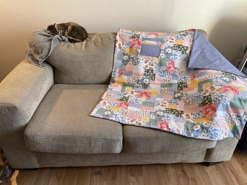 Puede incluir: Un sof&aacute; gris con una colcha de patchwork en tonos azules, rosas, amarillos y verdes. La colcha tiene un patr&oacute;n floral y est&aacute; doblada en el sof&aacute;.