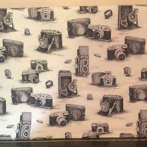 May include: A white fabric with a repeating pattern of black and white vintage cameras. The cameras are drawn in a realistic style and are arranged in a random pattern.