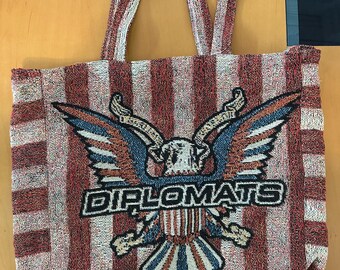 May include: A red and white striped tote bag with a woven design. The bag features an eagle emblem with outstretched wings, and the word "DIPLOMATS" is displayed below the eagle.