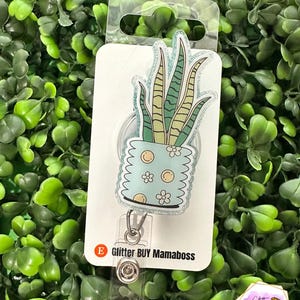 May include: A retractable badge reel featuring a potted snake plant design. The plant is in a light blue pot with white flowers. The reel has the text "Glitter BUY Mamaboss". The background is a bed of green leaves.