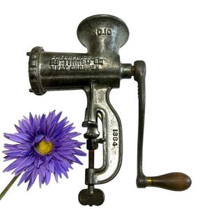 May include: A vintage cast iron Enterprise No. 10 meat chopper with a hand crank and wooden handle. The antique kitchen tool has a clamp for attaching to a table. A purple flower with a yellow centre is next to the chopper.