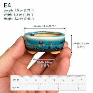 May include: A small oval ceramic bonsai pot with a blue and turquoise glaze featuring dark circular spots. The pot is labeled E4 and measures 1.77 inches in length, 1.30 inches in width, and 0.85 inches in height as indicated by the visible ruler.