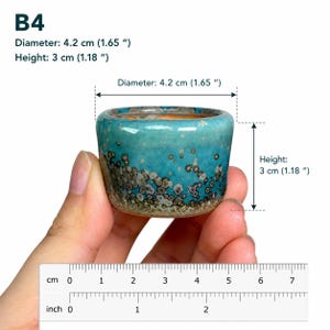 May include: A small, round ceramic pot labeled B4 with a turquoise and brown speckled glaze. The pot has a diameter of 1.65 inches and a height of 1.18 inches, as indicated by the text and the visible ruler in the image.