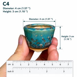 May include: A small ceramic cup with a teal and blue crystalline glaze featuring dark circular patterns. The cup is labeled C4 and measures 1.57 inches in diameter and 1.18 inches in height, as indicated by the text and visible ruler.