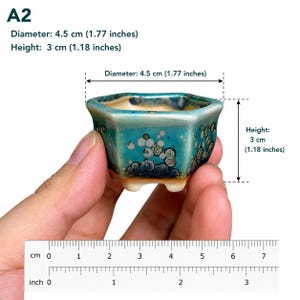 May include: A small hexagonal ceramic bonsai pot with a teal and blue crystalline glaze, featuring a beige interior and three short feet. The pot is labeled A2 and measures 1.77 inches in diameter and 1.18 inches in height as shown by a ruler.