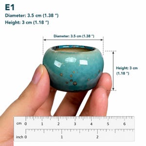 May include: A small, round, turquoise ceramic planter with a glossy finish and brown speckles. The planter is labeled E1 and measures 3.5 cm (1.38 inches) in diameter and 3 cm (1.18 inches) in height, as indicated by the visible ruler and text.