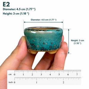 May include: A small, round, teal ceramic pot with a speckled glaze and three short feet. The pot has a diameter of 1.77 inches and a height of 1.18 inches, as indicated by the text and the visible ruler at the bottom of the image.