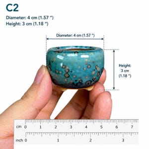 May include: A small, round, turquoise ceramic planter with a speckled glaze and three short feet. The planter is labeled C2 and measures 1.57 inches in diameter and 1.18 inches in height, as indicated by the text and visible ruler in the image.