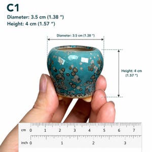 May include: A small, round, turquoise ceramic planter with a speckled, crystalline glaze pattern. The planter features three short feet at the base. Text indicates a diameter of 1.38 inches and a height of 1.57 inches, confirmed by a visible ruler.