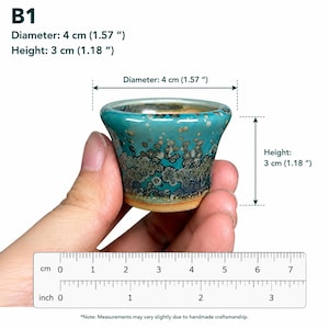 May include: A small ceramic pot with a teal and dark blue crystalline glaze, featuring a flared rim and a tan base. The pot is labeled B1 and measures 1.57 inches in diameter and 1.18 inches in height, as indicated by the visible ruler.