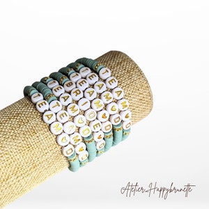 Puede incluir: Una pila de diez pulseras con cuentas blancas y letras doradas. Las pulseras deletrean las palabras "I AM", "TEAM", "MADE", "LOVE", "DREAM", "BRIAR", "TEA" y "MOM".