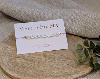 Op de afbeelding: Een lichtpaarse koordarmband met witte letterkralen die het woord MARRAINE vormen, geflankeerd door twee goudkleurige kralen. De armband is bevestigd aan een witte kaart met de tekst Veux tu être MA MARRAINE, Créatrice de bijoux faits mains, Made in France.