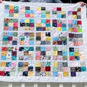 May include: A handmade quilt with a patchwork design. The quilt features a grid of colorful fabric squares in various patterns and hues, including red, blue, yellow, and green, set against a white background. The quilt is held up by a person.